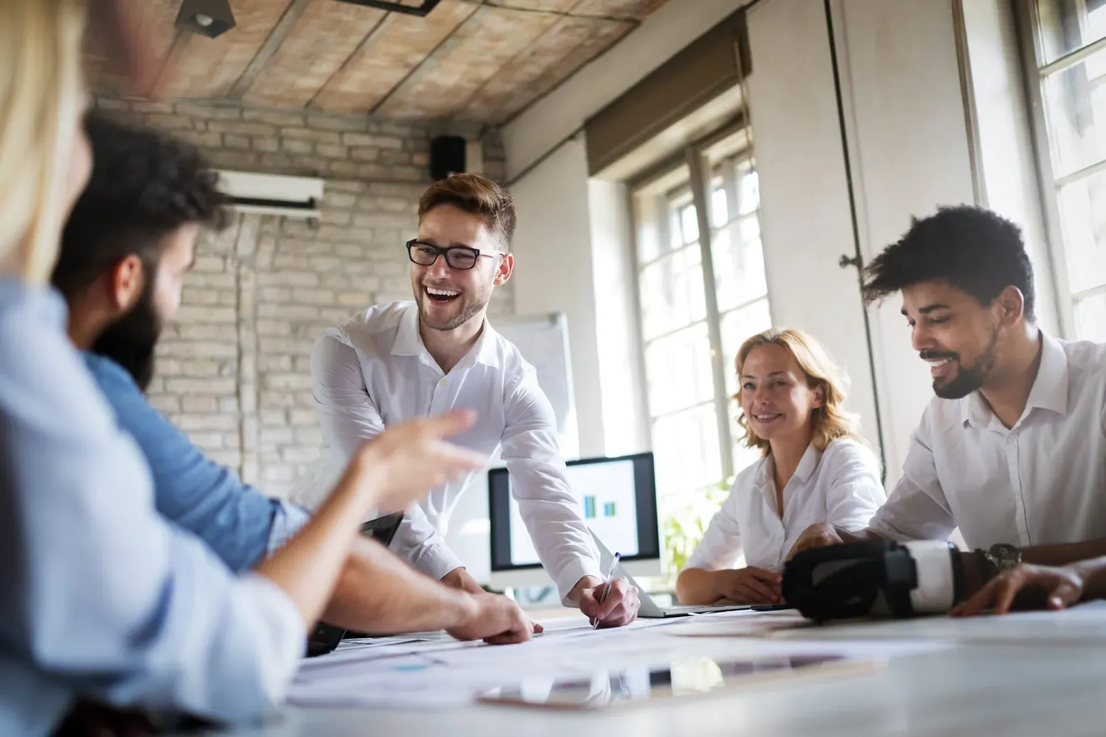 Team laughing together around a table of papers in a brick-walled office meeting room