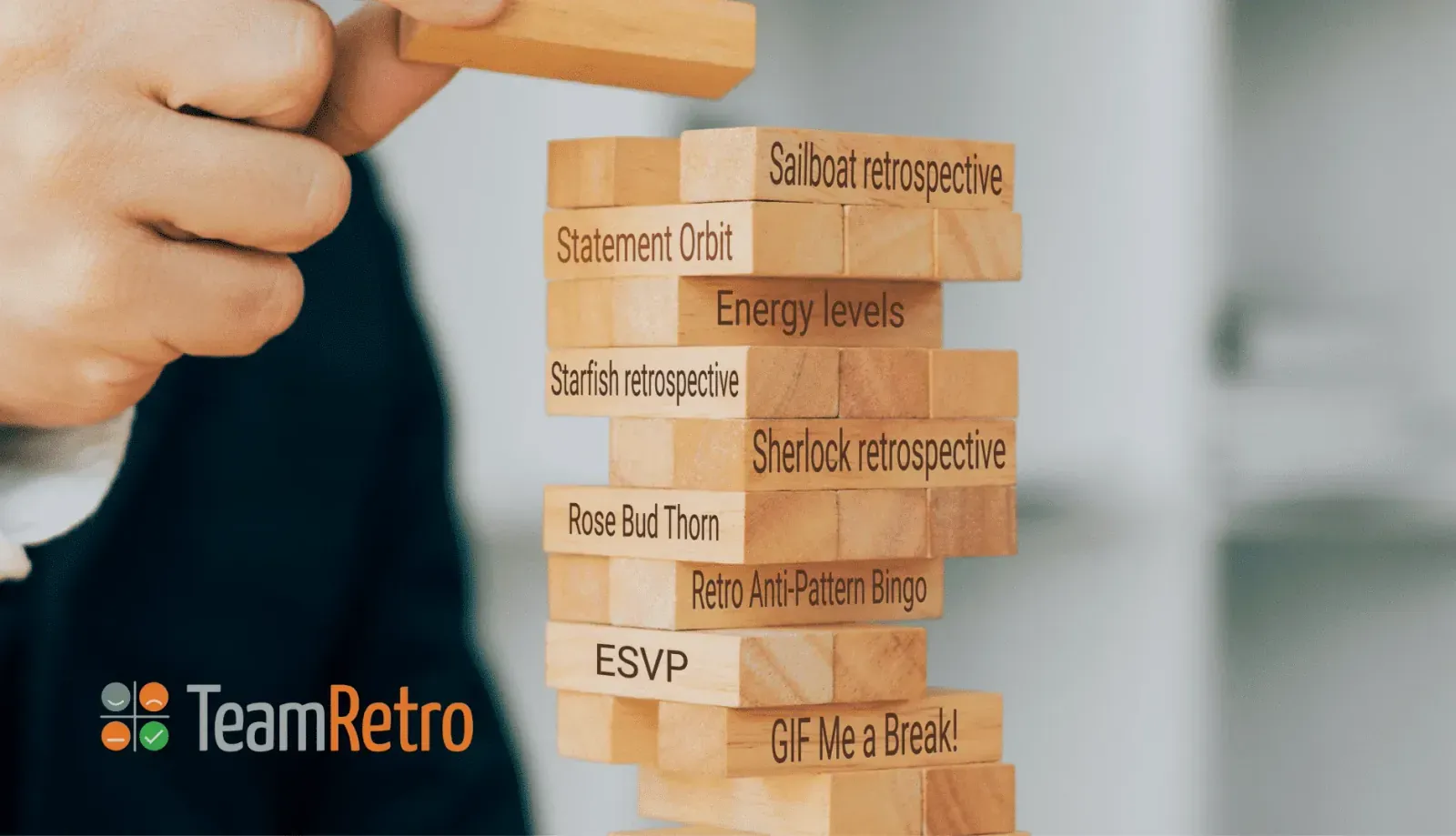 Hand stacking a wooden block tower labeled with retro game names such as Sailboat, ESVP, Statement Orbit and Anti-Pattern Bingo
