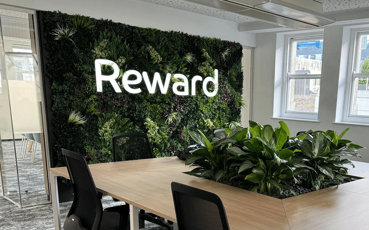 Reward office with an illuminated Reward sign on a green plant wall above shared desks and a planter