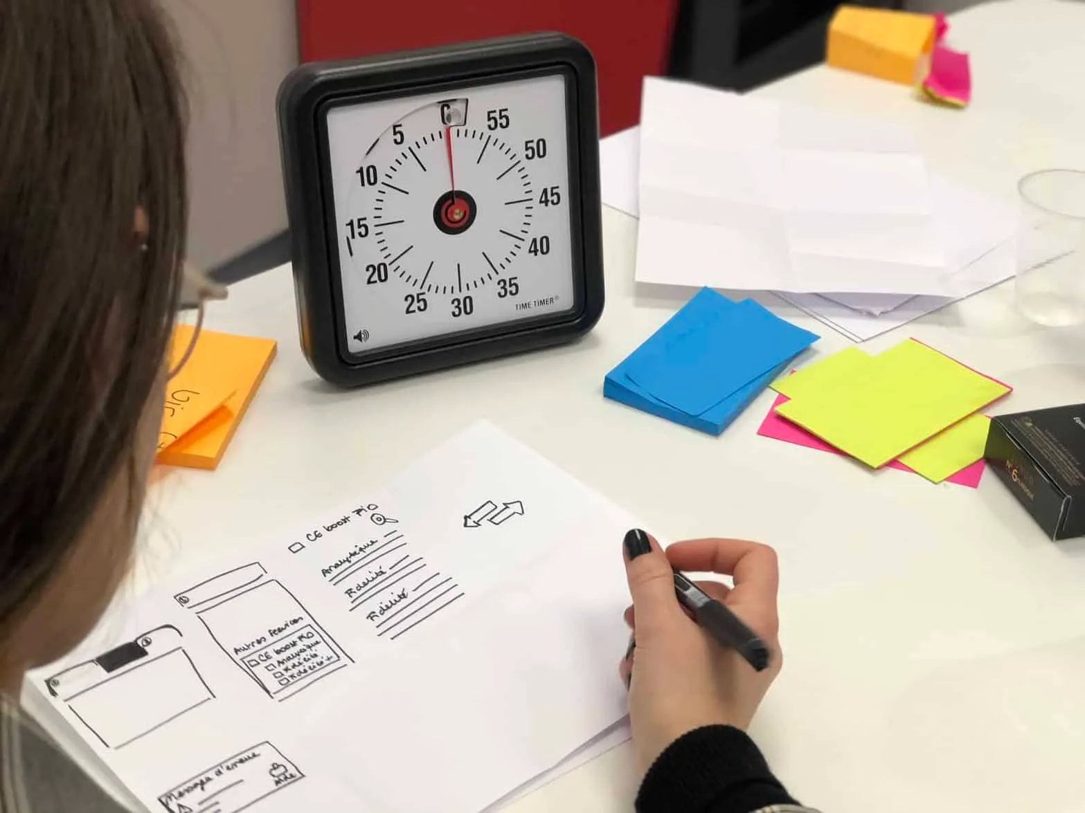 Person sketching wireframes at a desk beside a Time Timer countdown clock and colored sticky notes